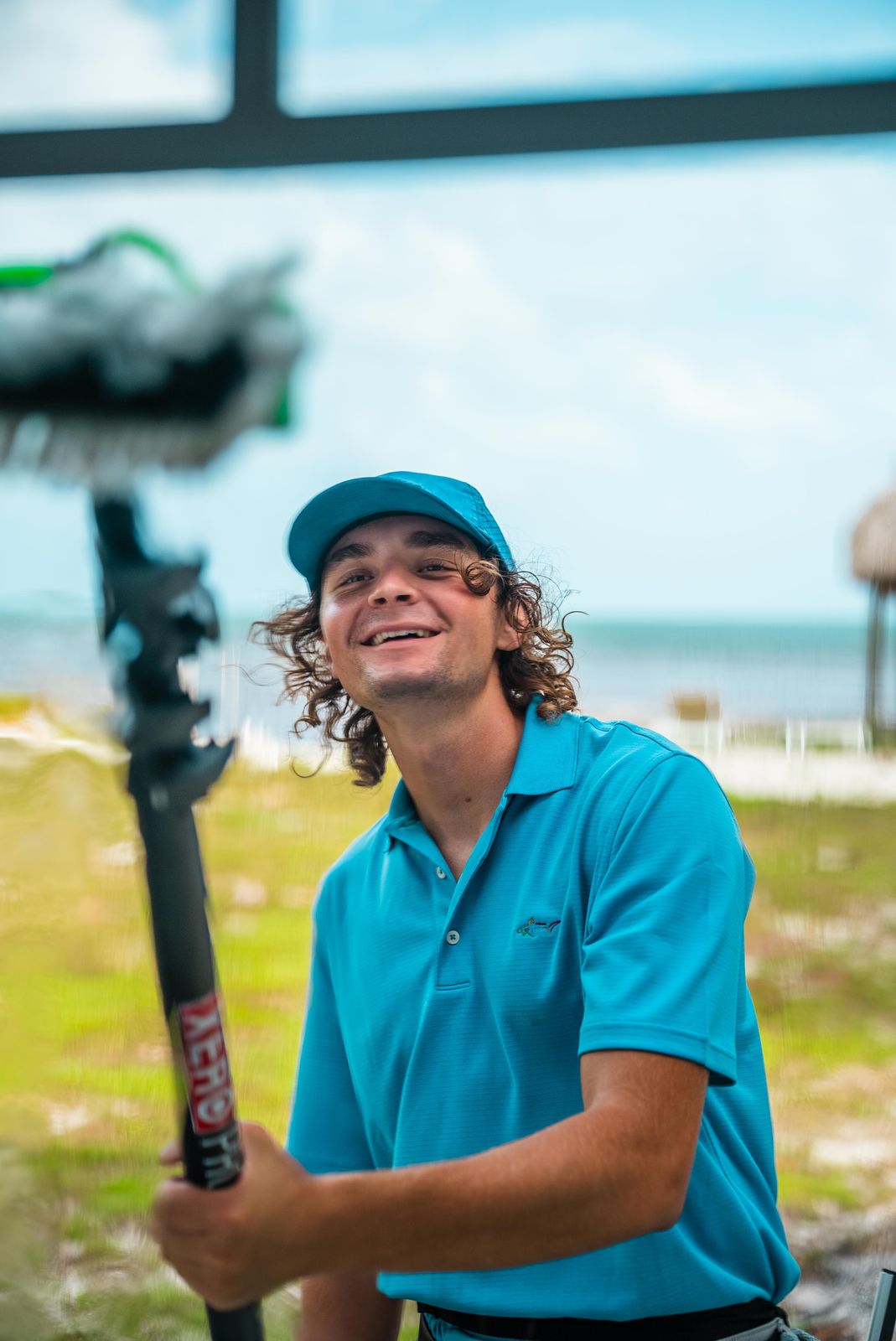 Sea Clear window cleaner smiling through freshly washed glass, water-fed brush in hand, with a tiki hut and the ocean behind him