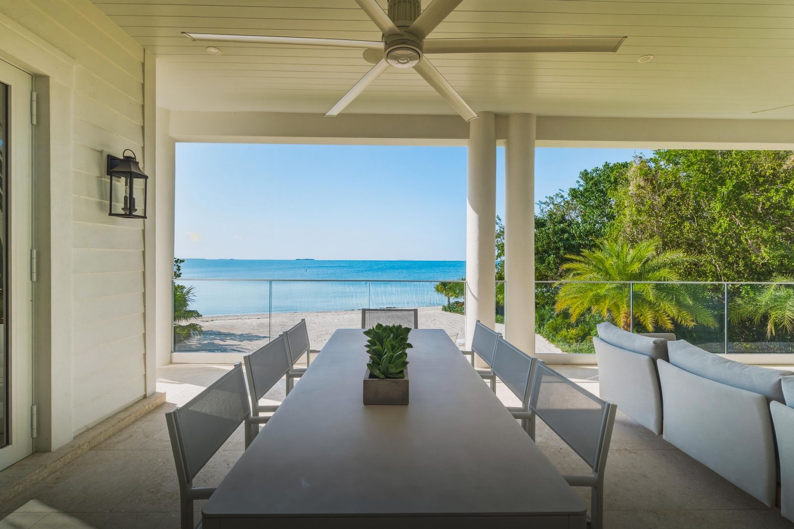 Covered waterfront porch in the Florida Keys with a long outdoor dining table, palms and calm turquoise water beyond the glass railing