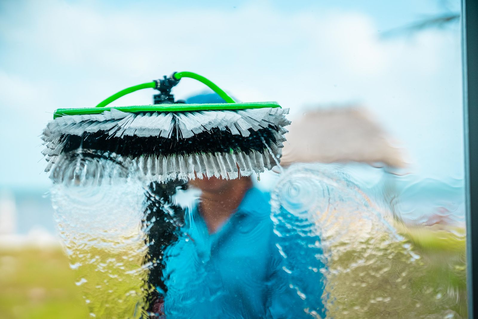 Water-fed brush washing a window, seen from inside with a tiki hut and the ocean beyond